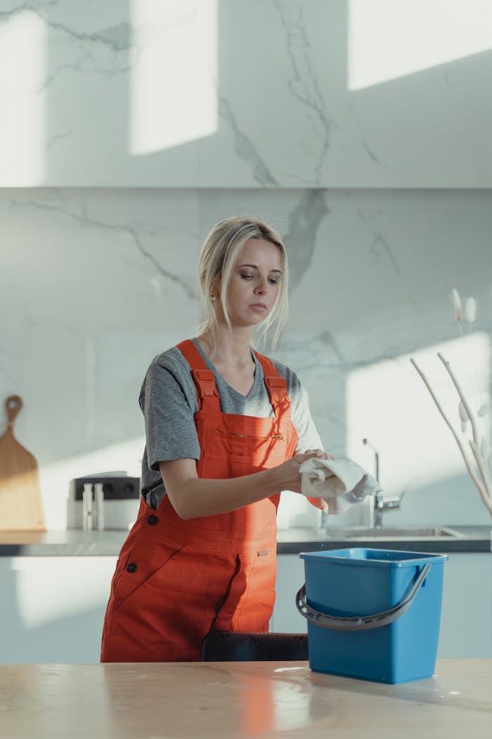 about-us-01 A woman wearing orange overalls cleans a kitchen counter with a rag and pail in a modern kitchen.
