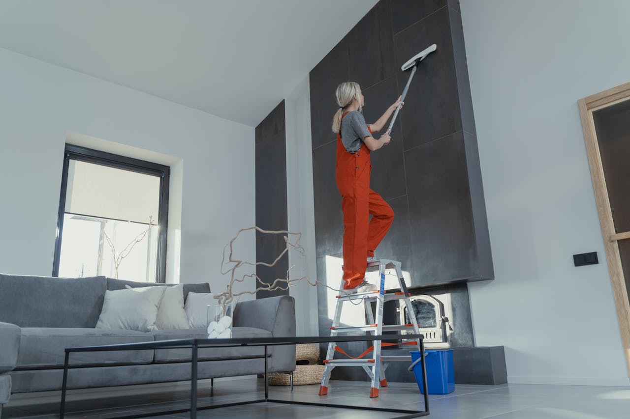 heros-img A woman stands on a ladder in a modern living room, cleaning the high wall with a mop.